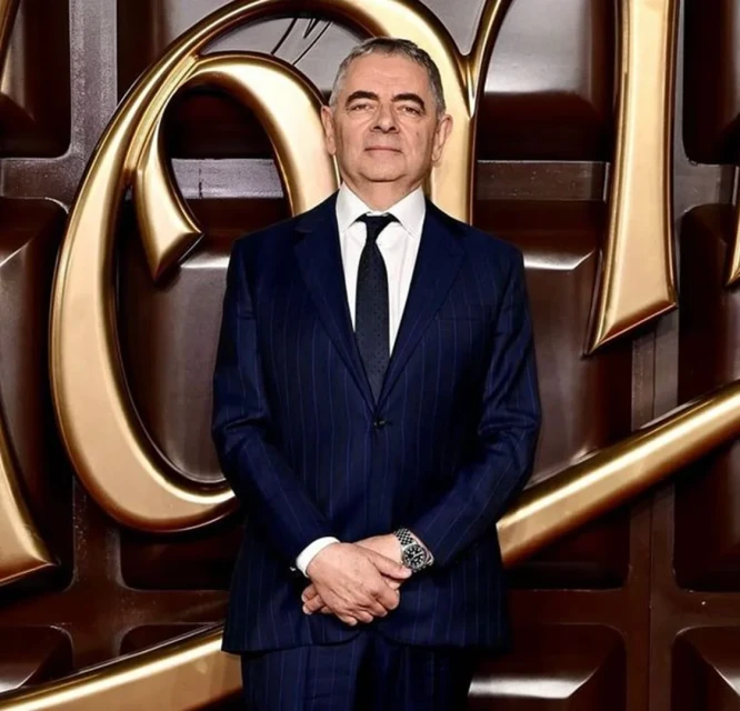 Headshot of adult male wearing a pinstripe suit, standing in front of a dark brown and gold background with abstract chocolate-like shapes.