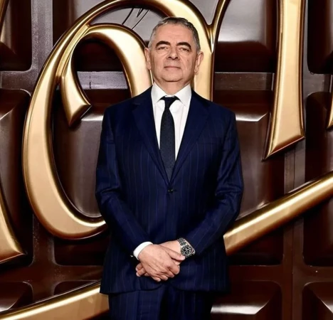 Headshot of adult male wearing a pinstripe suit, standing in front of a dark brown and gold background with abstract chocolate-like shapes.
