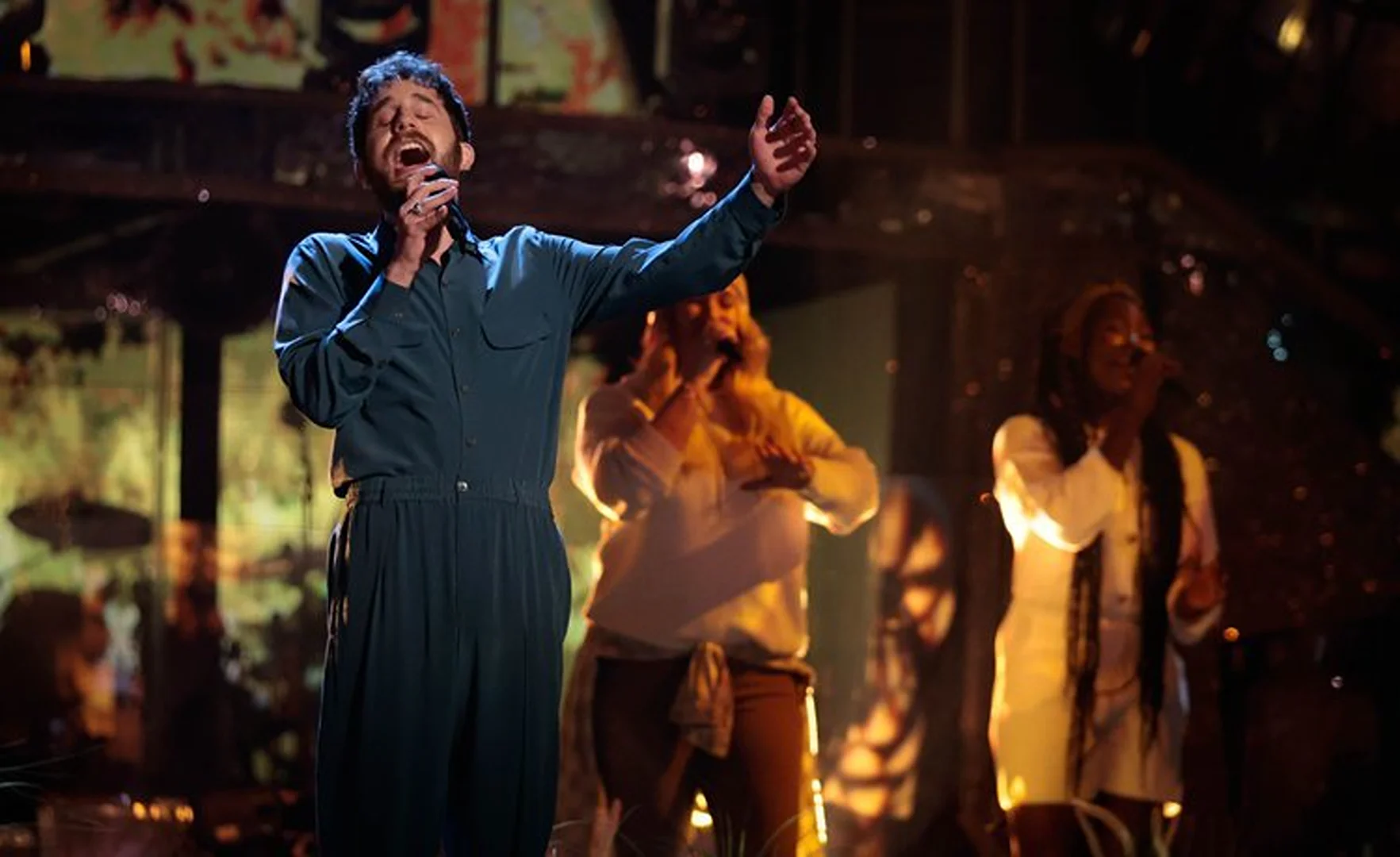 Male vocalist performs on stage with two female backing singers in a concert setting, illuminated by warm lighting and stage effects.