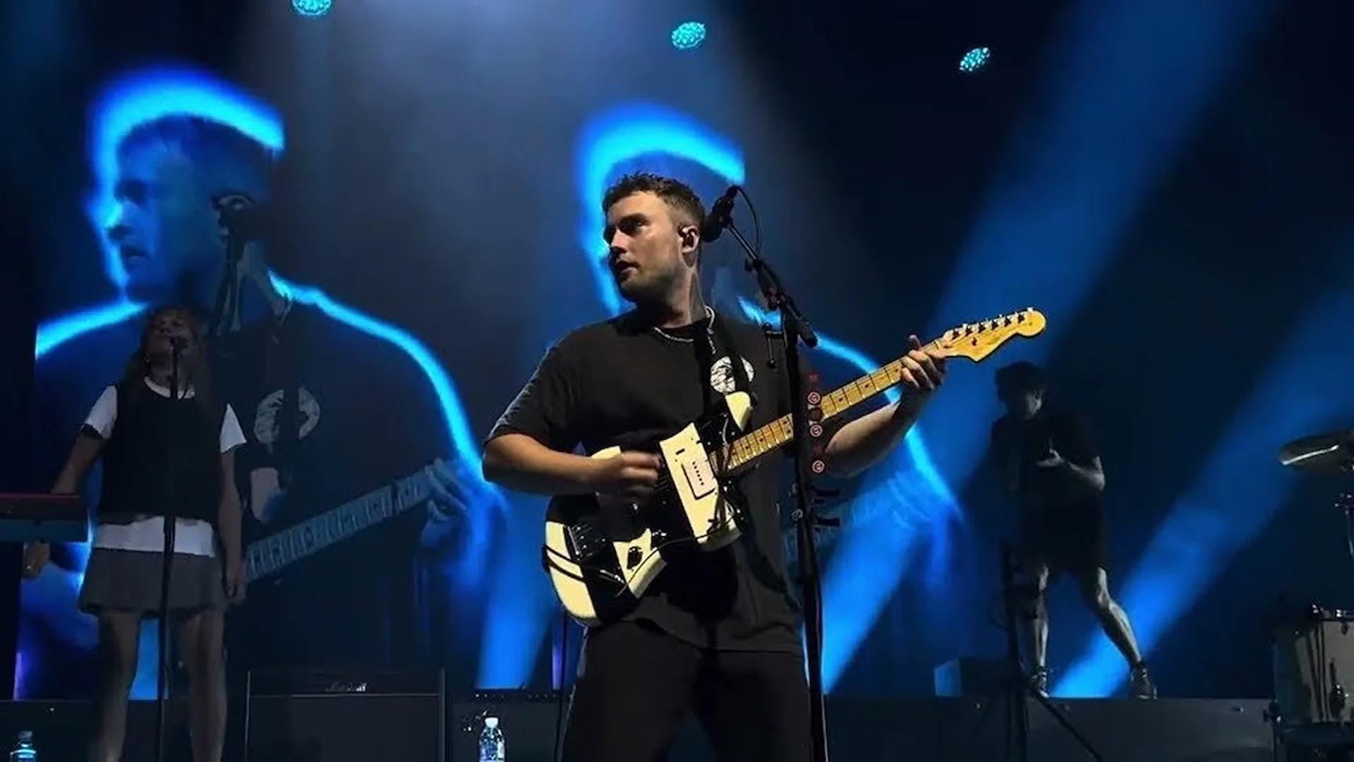Musician performing live on stage with a white electric guitar under bright blue concert lighting; other band members visible.