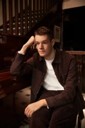 A young man in a dark brown corduroy jacket and white t-shirt resting his head on his hand indoors.