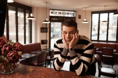 Man wearing a black and white striped sweater leaning on a table inside a dimly lit café with pendant lamps.