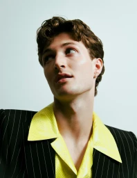 Close-up portrait of a man with dark curly hair wearing a pinstripe jacket and yellow collared shirt against a pale background