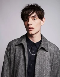 Close-up portrait of a young man wearing a grey tweed jacket over a dark patterned shirt and a silver chain necklace.