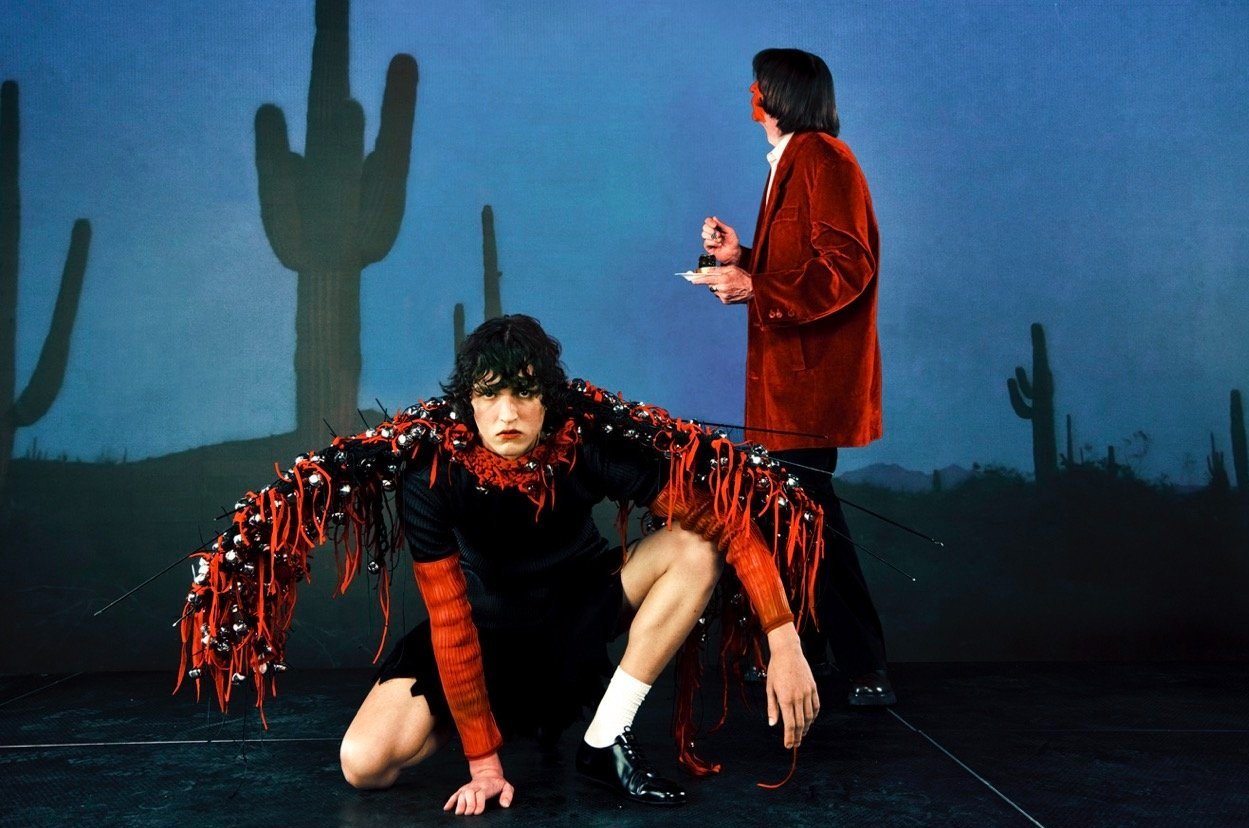 Two people in a studio setting with a desert backdrop. The person in the foreground is kneeling and wearing a unique costume.