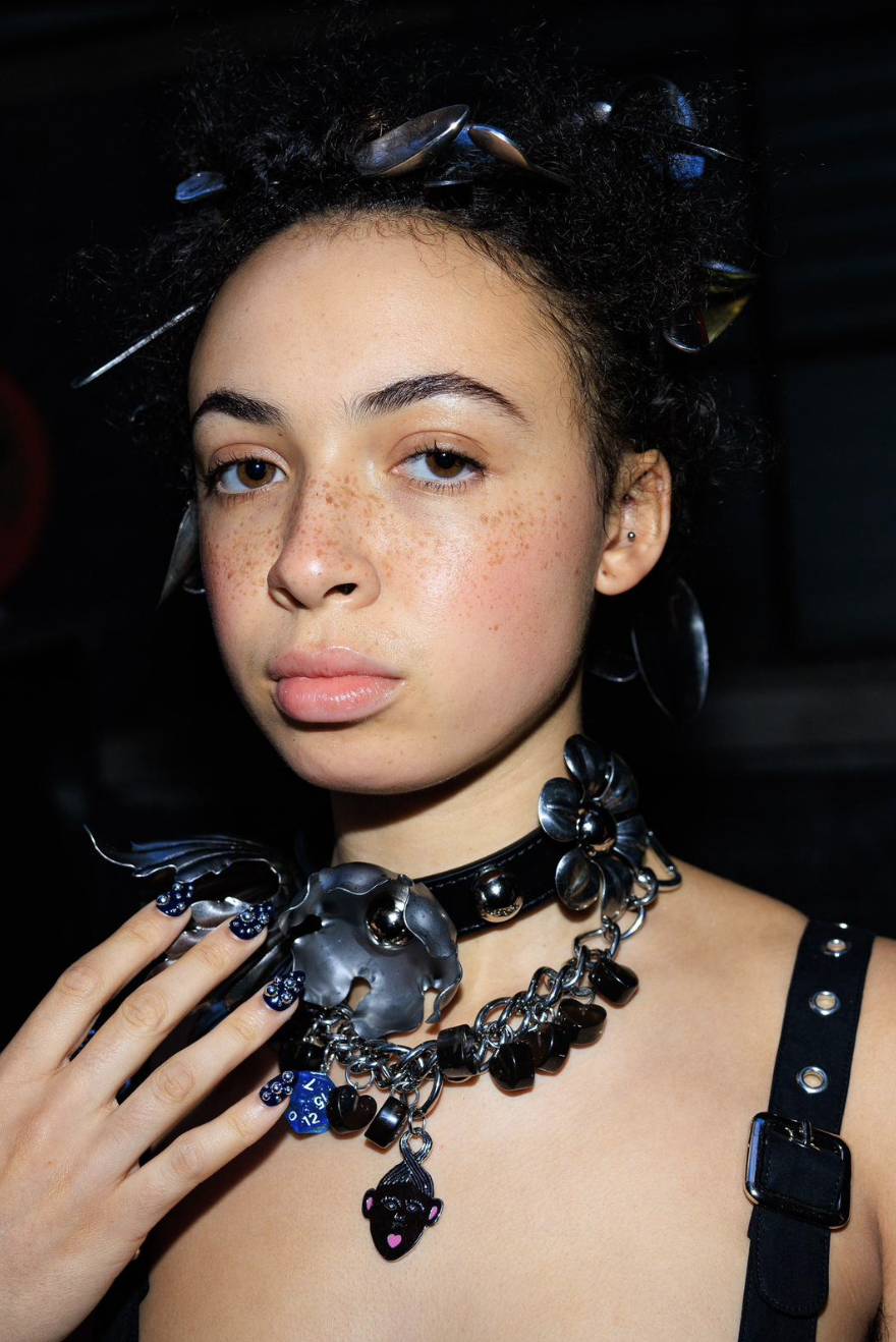 Close-up shot of an adult female with dark, curly hair and a heavy, black and silver jeweled collar necklace in a dark indoor environment.