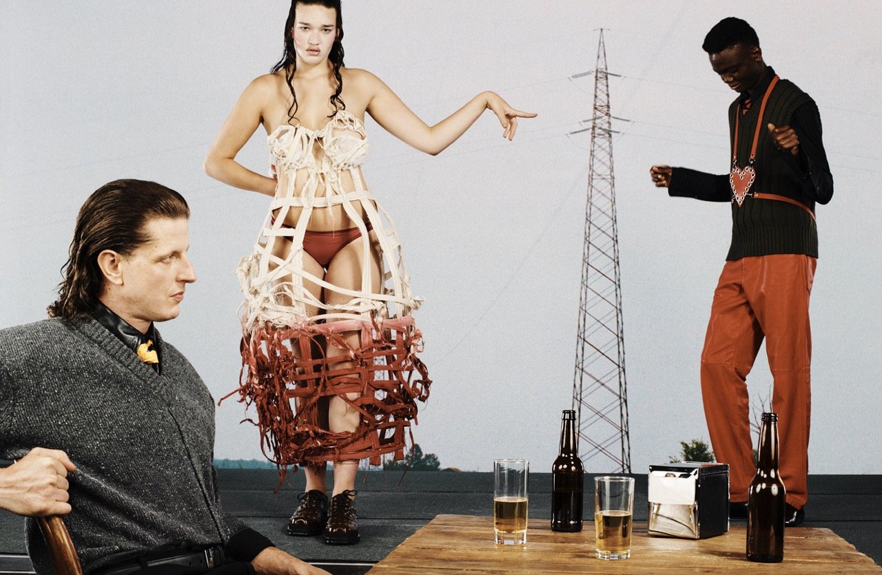 Fashion shoot featuring three people: a seated man, a woman in an unusual dress, and a man in rust-colored pants, set against an outdoor backdrop.
