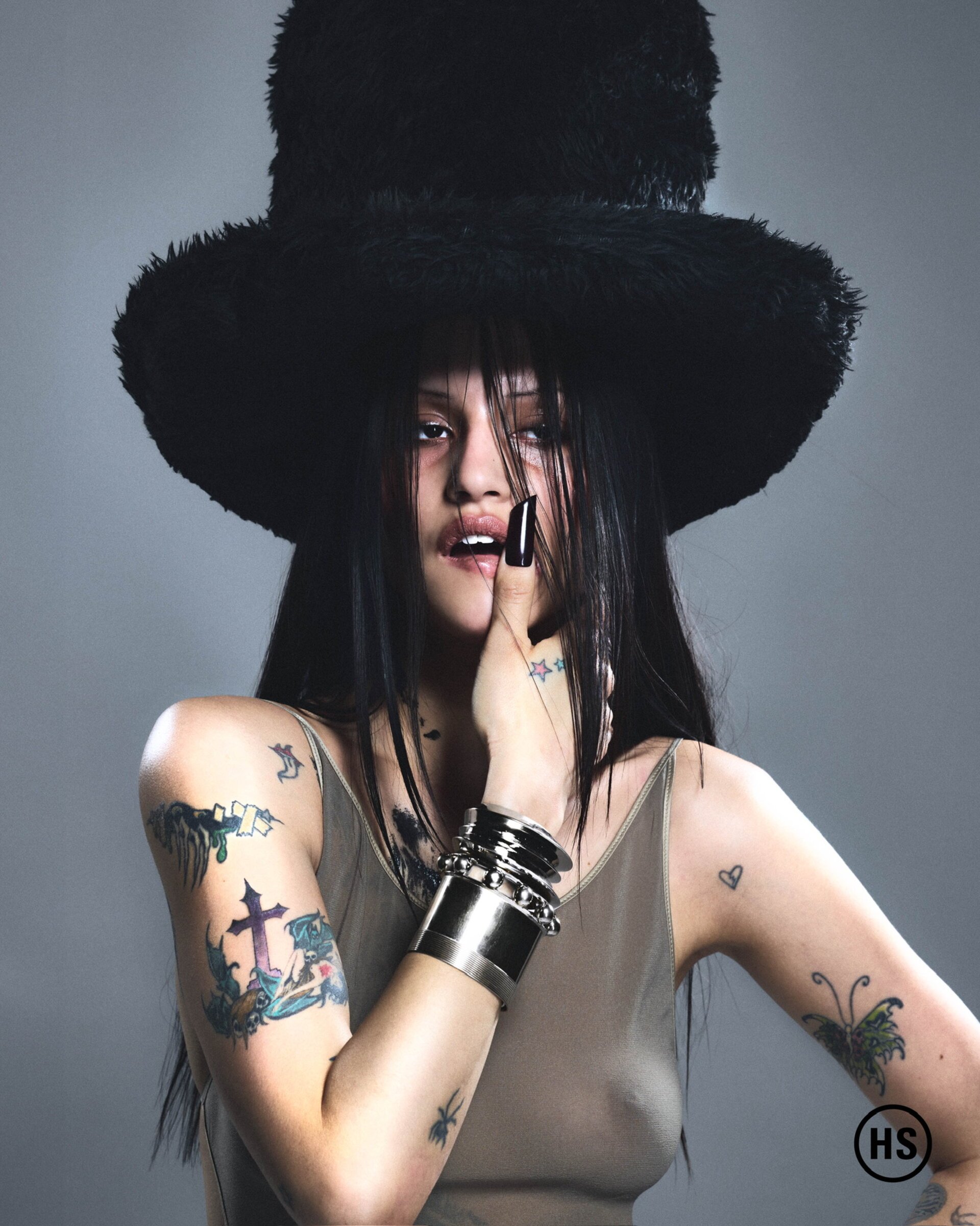 Portrait of a woman with long black hair and tattoos, wearing a black hat and multiple silver bracelets, against a grey backdrop.
