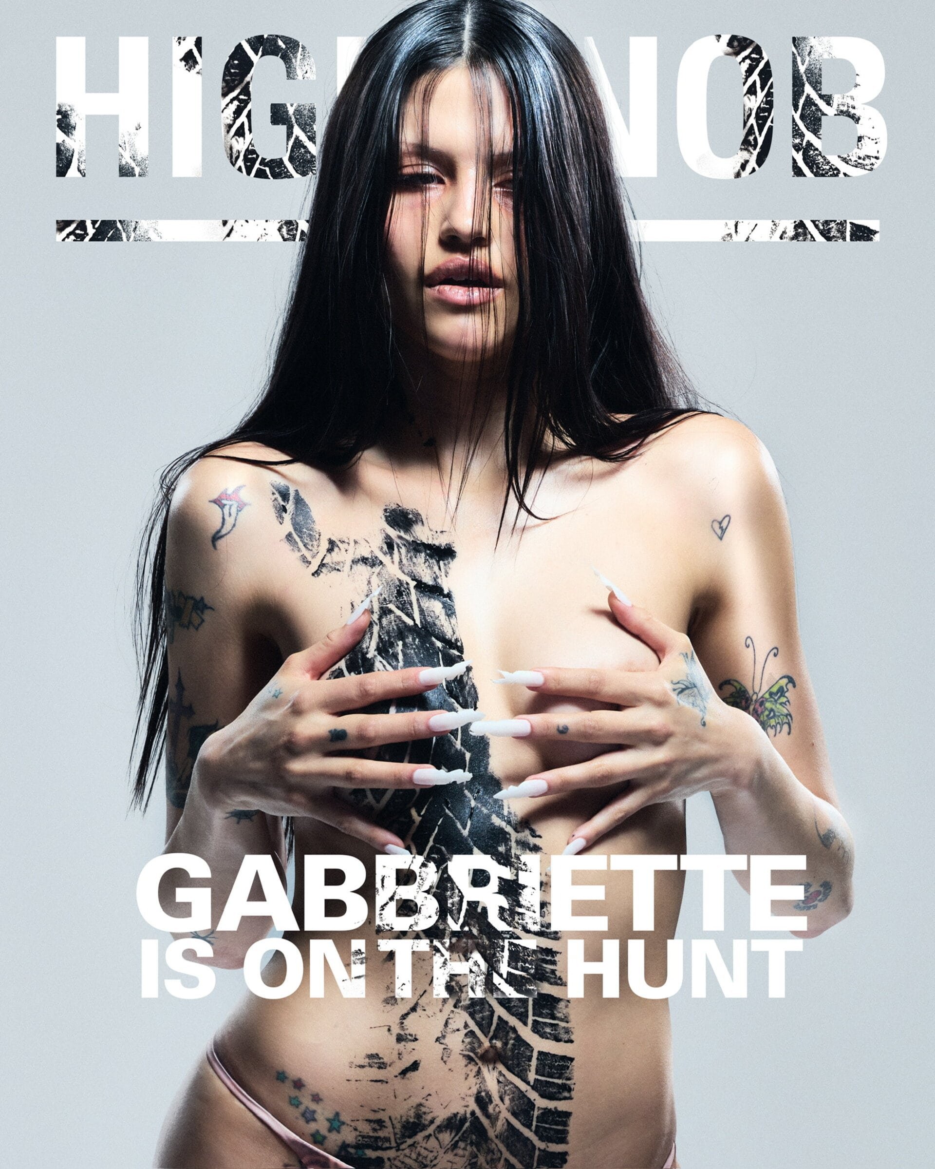 Woman posing for a magazine cover with body paint and tattoos on a light grey background. The cover text is visible above the subject.