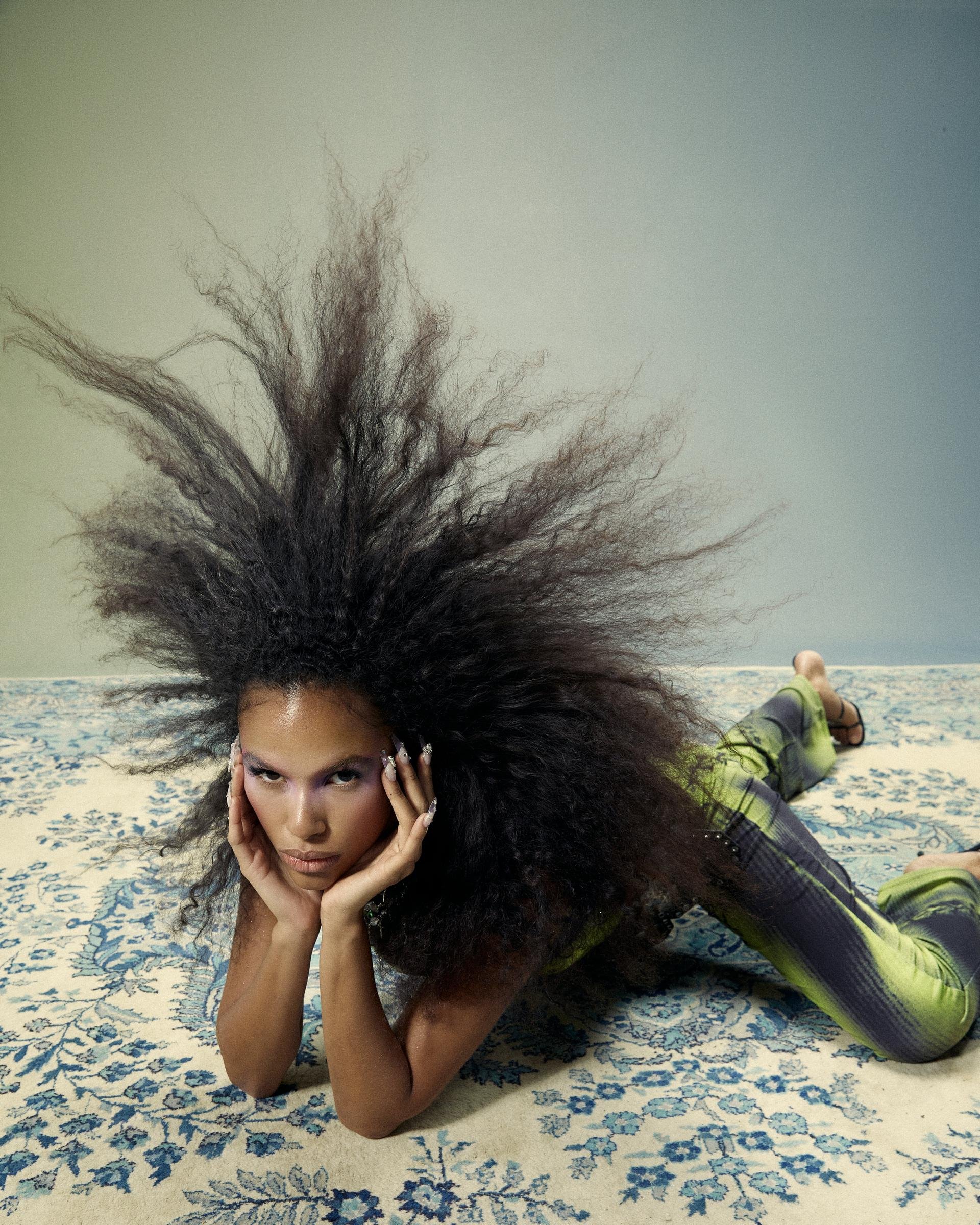 Elegant woman with an afro lying on a floral patterned rug. She is wearing a tight fitting green outfit with a light blue background.