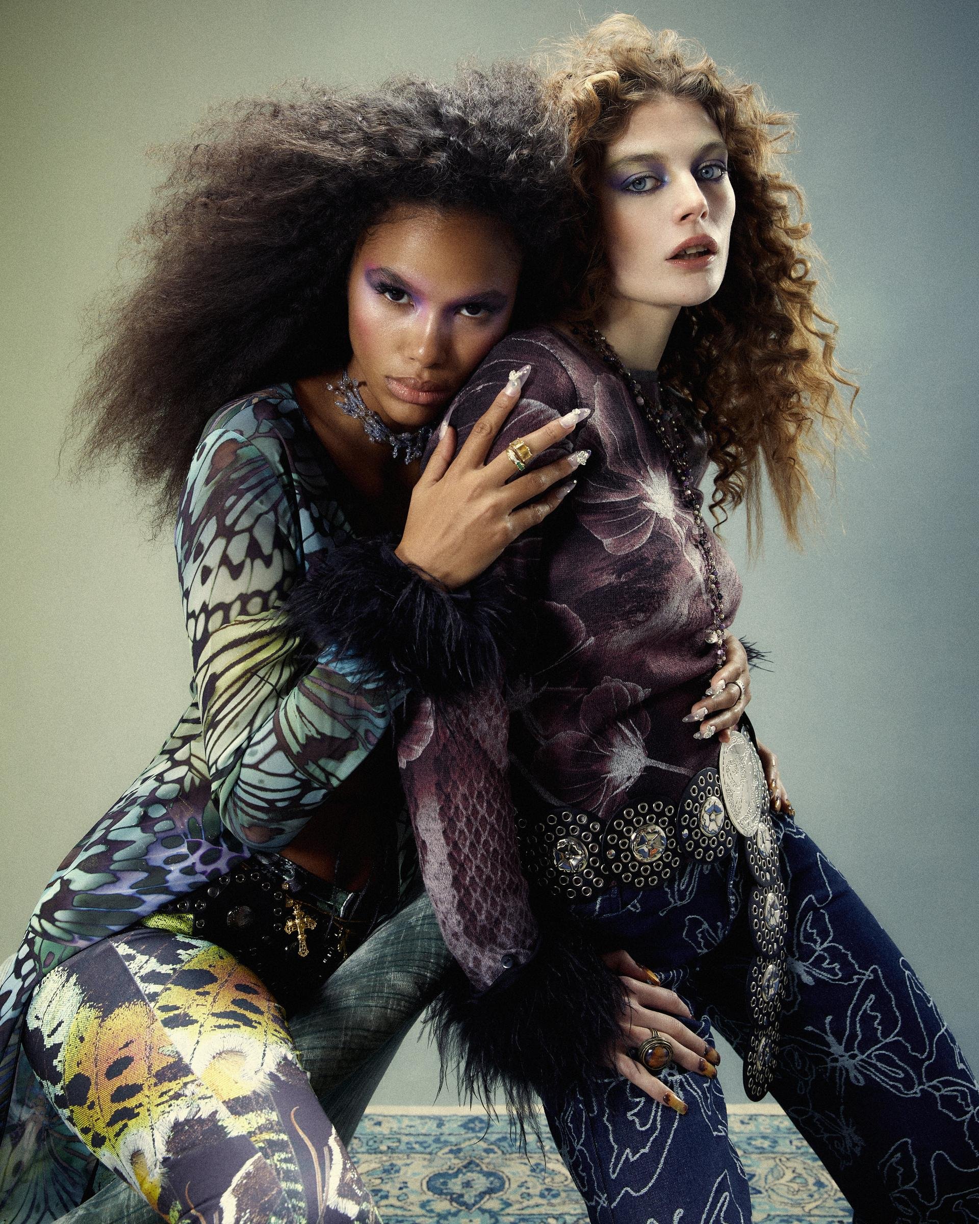 Two fashion models in patterned tops and bottoms pose indoors with eye makeup. One model is Black with an afro hairstyle and the other is white with red hair.