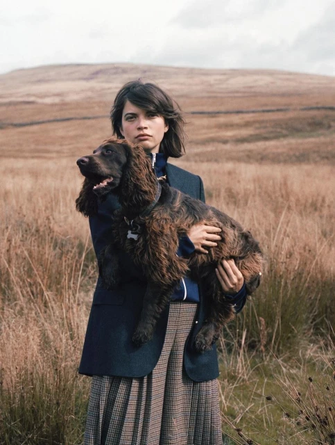 Young woman in blazer and pleated skirt holding brown Cocker Spaniel dog in a rural landscape of golden grass and rolling hills.