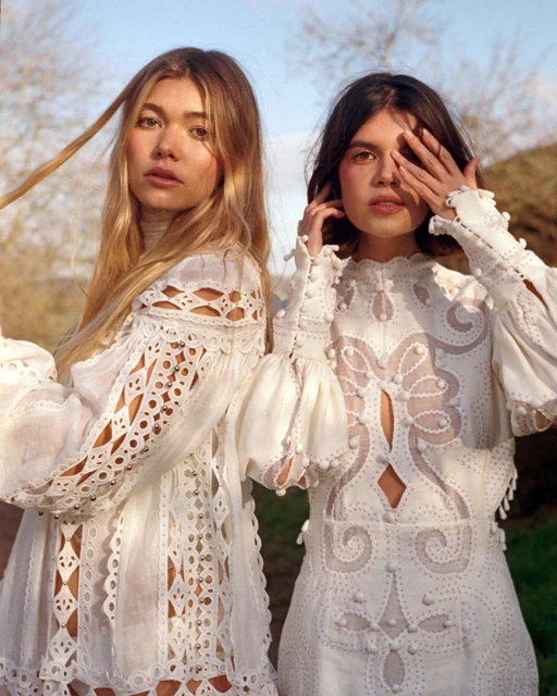 Two fashion models wear white, lacy dresses outdoors with a shallow depth of field. Light is diffused with natural sun illuminating the shot.