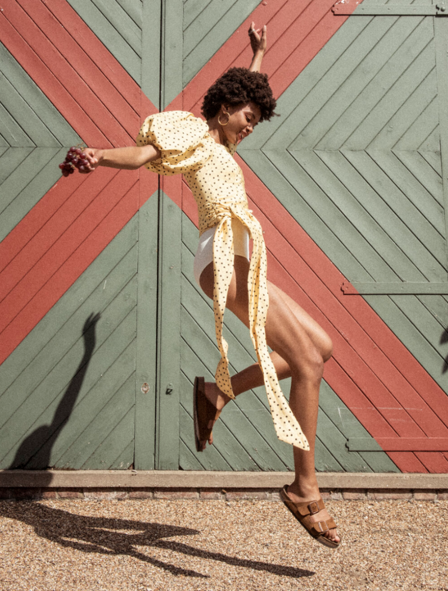 Woman with afro jumps happily in yellow polka dot top, white shorts, and brown sandals in front of barn doors, bright sunlight.