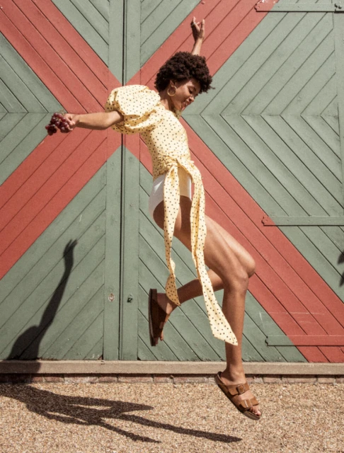 Woman with afro jumps happily in yellow polka dot top, white shorts, and brown sandals in front of barn doors, bright sunlight.