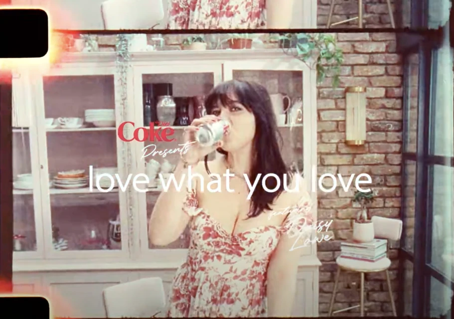 Woman drinks from a Coca-Cola can in an indoor setting with a cabinet behind her. A brick wall and window add background details.