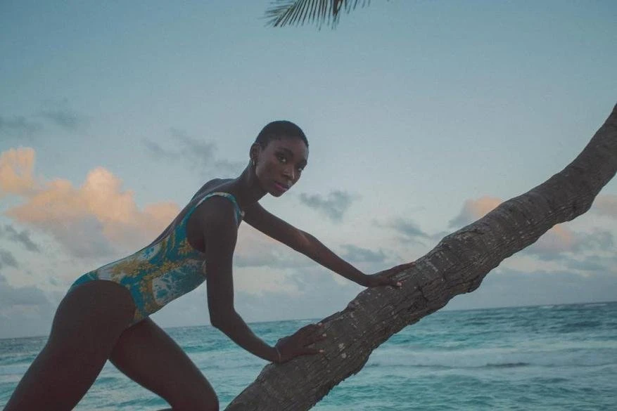 Adult woman in patterned swimsuit posing on a palm tree at a beach with ocean background, under a partly cloudy sky, low angle.