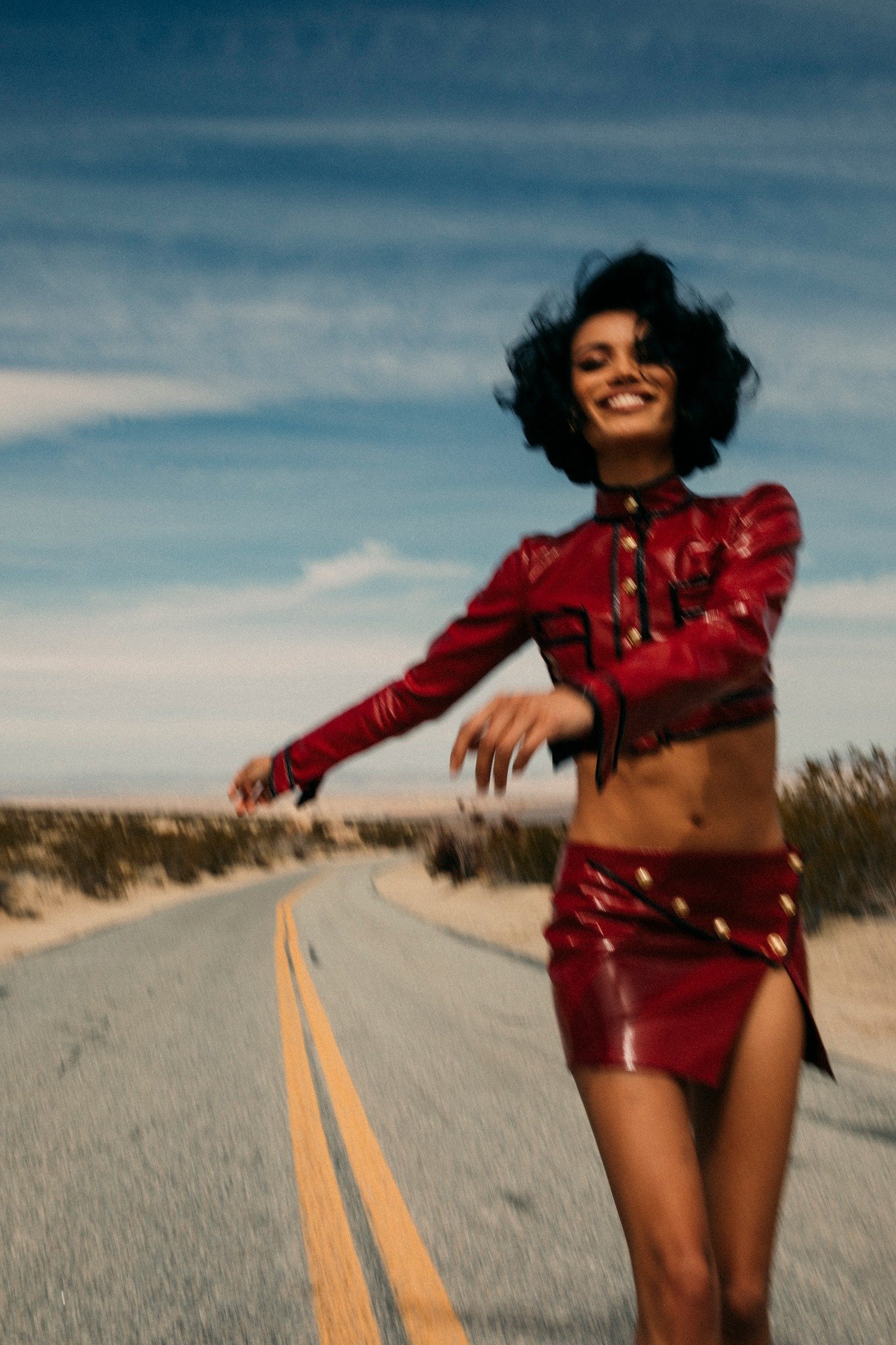 A smiling woman walks on a desert road wearing a red patent leather cropped jacket and matching mini skirt under a bright blue sky.