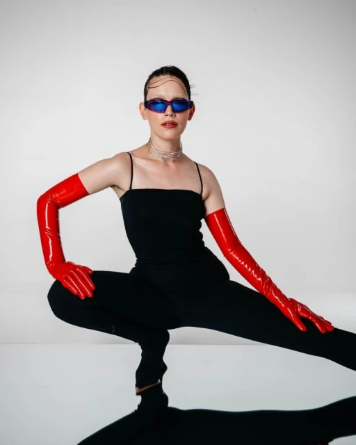 Fashionable woman in black bodysuit and red gloves poses in a photo studio with cool blue sunglasses and reflective floor, full shot.