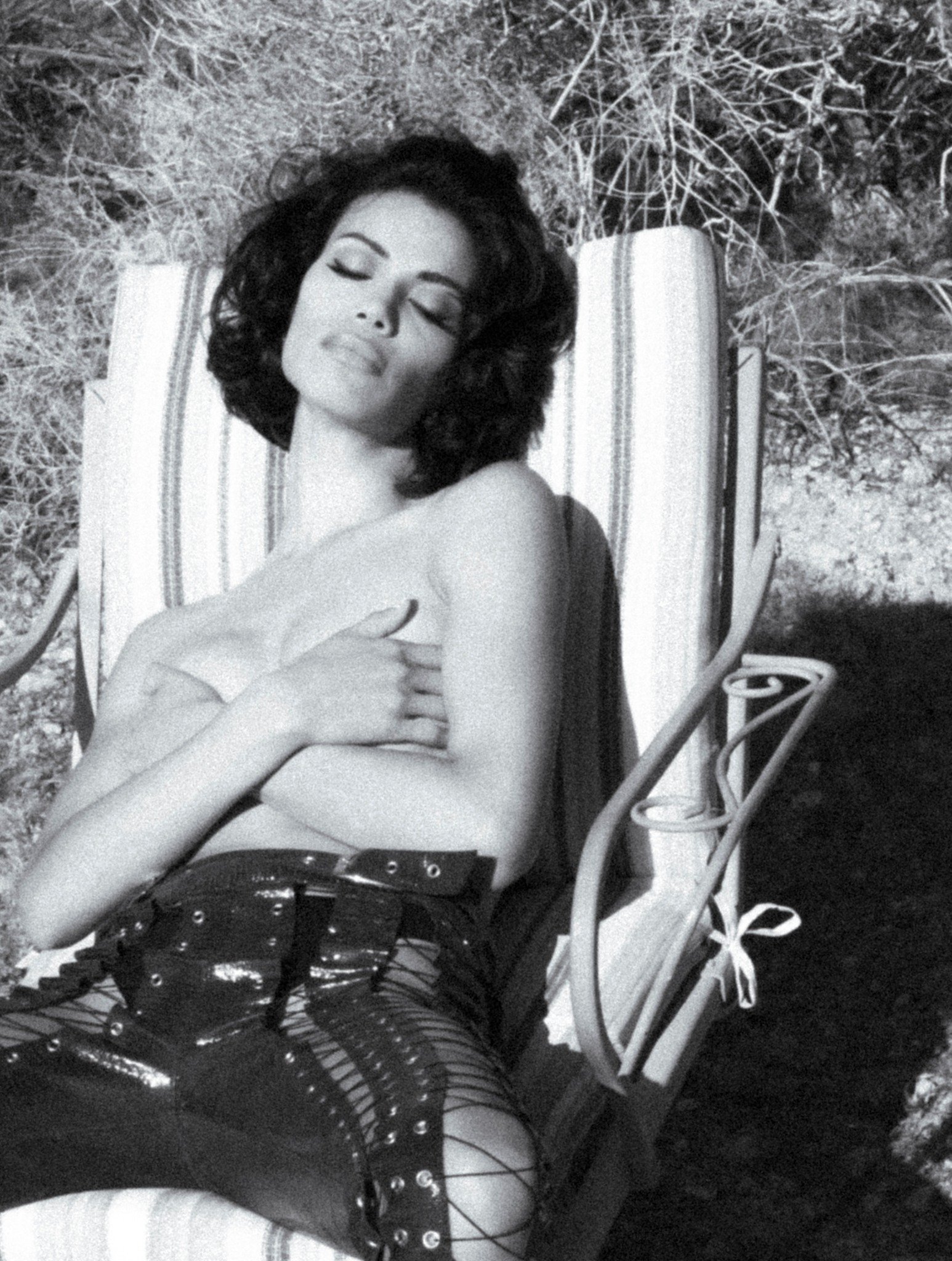 Monochrome image of a woman reclining on a striped lawn chair, facing upward with her eyes closed, wearing dark, laced-up pants.