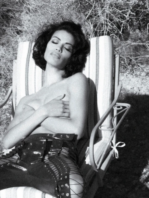 Monochrome image of a woman reclining on a striped lawn chair, facing upward with her eyes closed, wearing dark, laced-up pants.