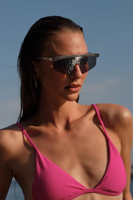 A woman with wet hair wears sunglasses and a pink bikini top against a blurred-blue sky background. Focus on fashion and summer style.