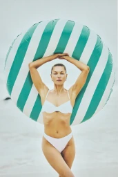 Woman in white bikini holding a large green and white striped inflatable ring above her head on a sandy beach.