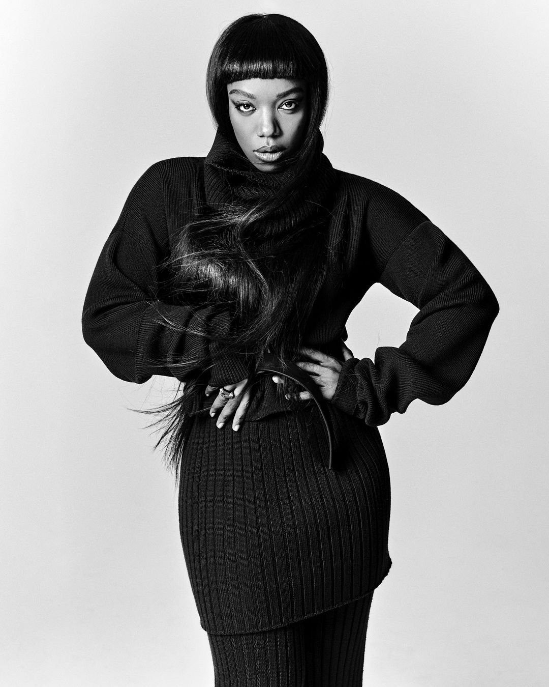 Black and white portrait of a woman with long dark hair wearing a black turtleneck sweater and skirt, hands on her hips.
