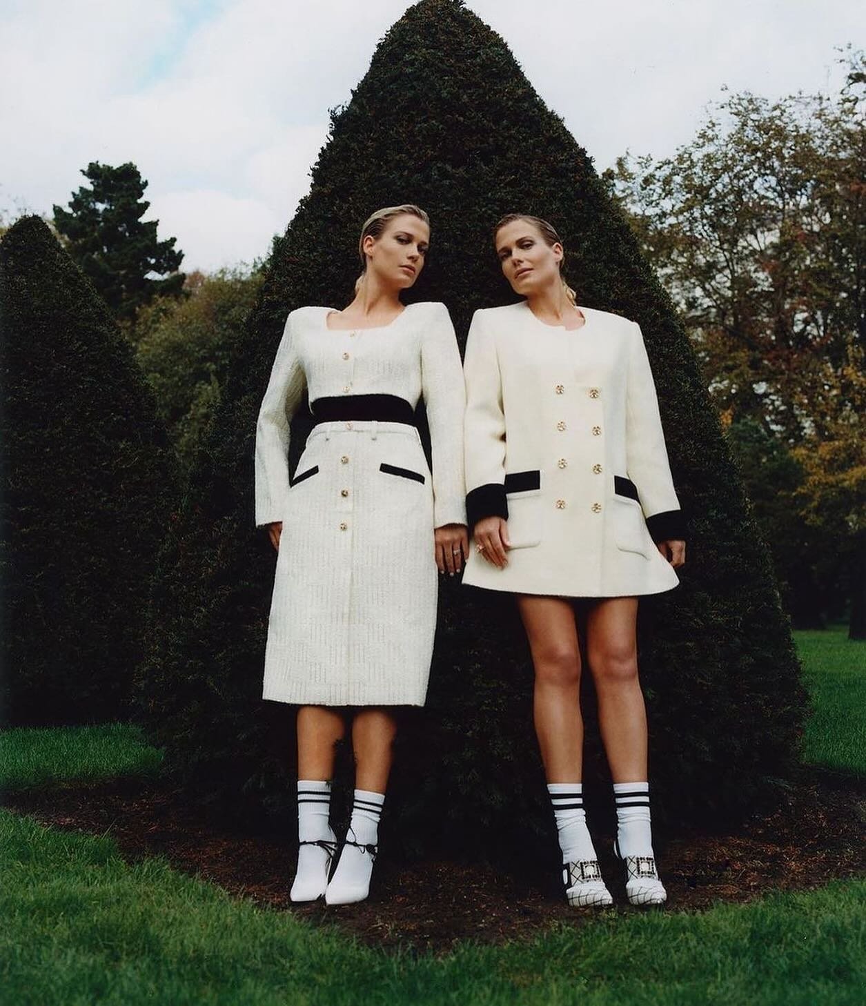 Two fair-skinned women pose outdoors in white and black trimmed fashion dresses, accessorized with white heels and socks, amidst lush greenery.