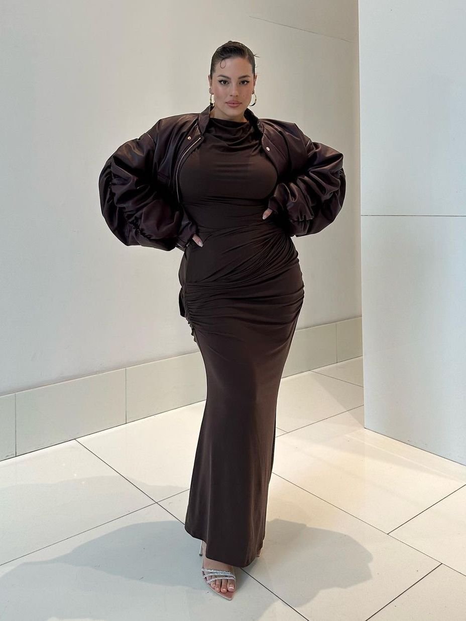 Ashley Graham poses in a brown dress and jacket against a neutral wall, smiling, and wearing hoop earrings and sandals.