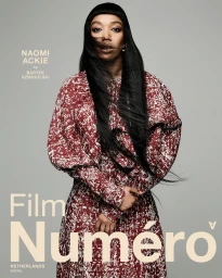 Portrait of actress Naomi Ackie wearing a red patterned dress against a plain grey studio backdrop.