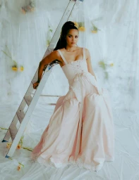 Woman in a pale pink corset gown sitting next to a metal ladder against a crinkled plastic backdrop with yellow flowers.