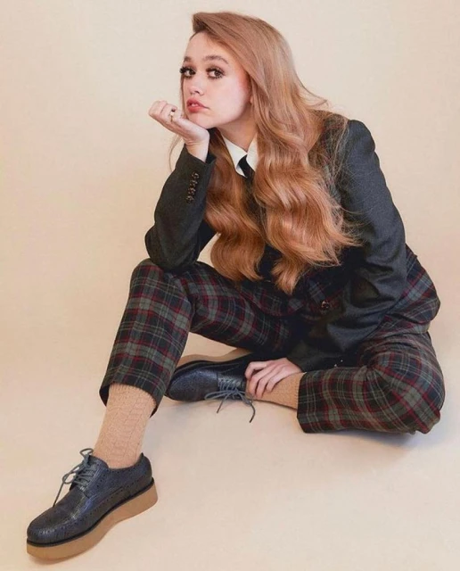 A red-haired woman in a dark-grey plaid suit sits in a photo studio with a neutral background, looking at the camera.