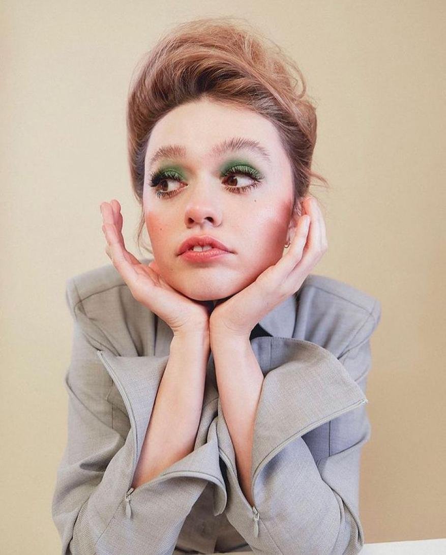 Close-up portrait of a young adult woman with green eye shadow, posing with her hands near her face against a beige background.