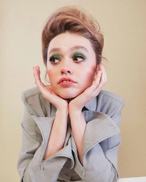 Close-up portrait of a young adult woman with green eye shadow, posing with her hands near her face against a beige background.