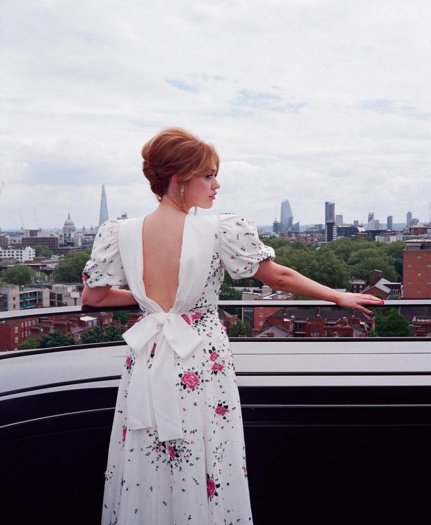 Woman in a white floral open-back dress poses on a rooftop with a city skyline and cloudy sky in the background in an outdoor shot.