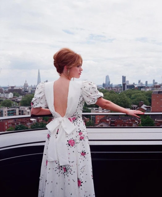 Woman in a white floral open-back dress poses on a rooftop with a city skyline and cloudy sky in the background in an outdoor shot.