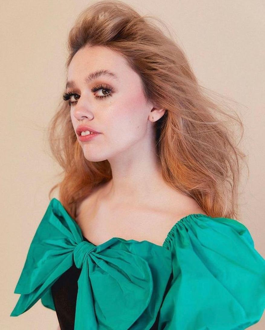 Portrait of a woman with blonde-reddish hair wearing a green bow dress, posing in front of a light beige backdrop, studio lighting.