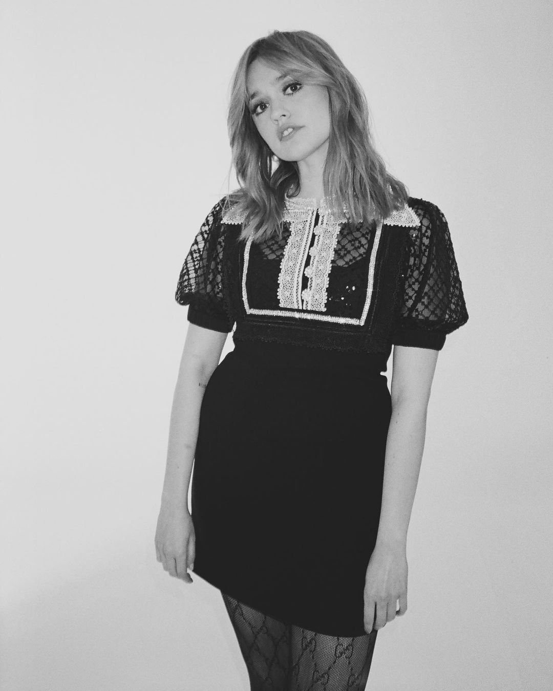 Monochrome portrait of a woman in a black dress with lace details, medium brown hair and light skin, posing against a bright backdrop.