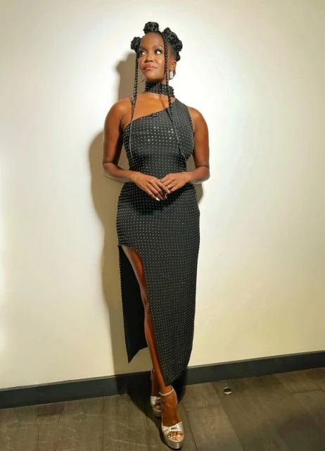 Woman in a black one-shoulder gown with hair styled in knots, posing against a neutral wall, accessorized with silver heels and dangling earrings.