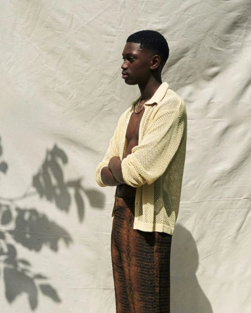Portrait of a young, dark-skinned man in a casual sweater and brown trousers posing in front of a white backdrop with tree shadows.