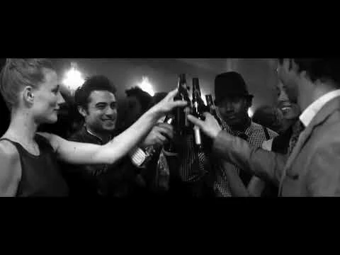 Monochrome image captures a group clinking beer bottles at an event, with smiling faces and formal attire, in an indoor setting.