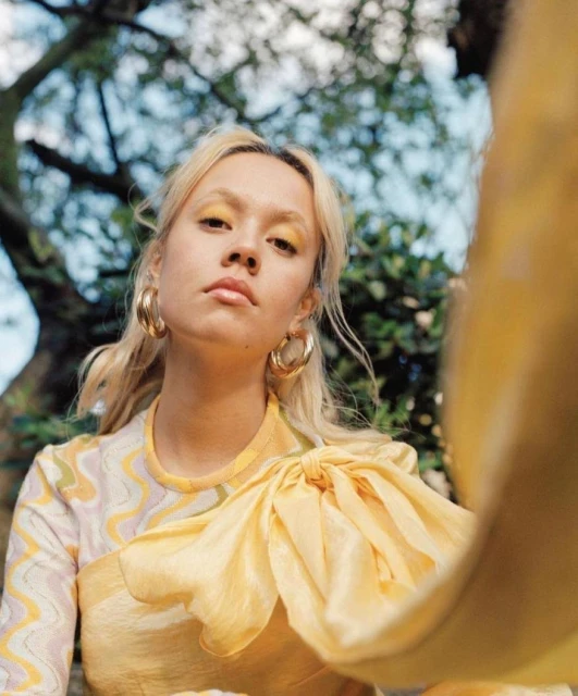 Portrait of a young woman with blonde hair and yellow eyeshadow in an outdoor setting with trees and soft lighting. Fashion shot.