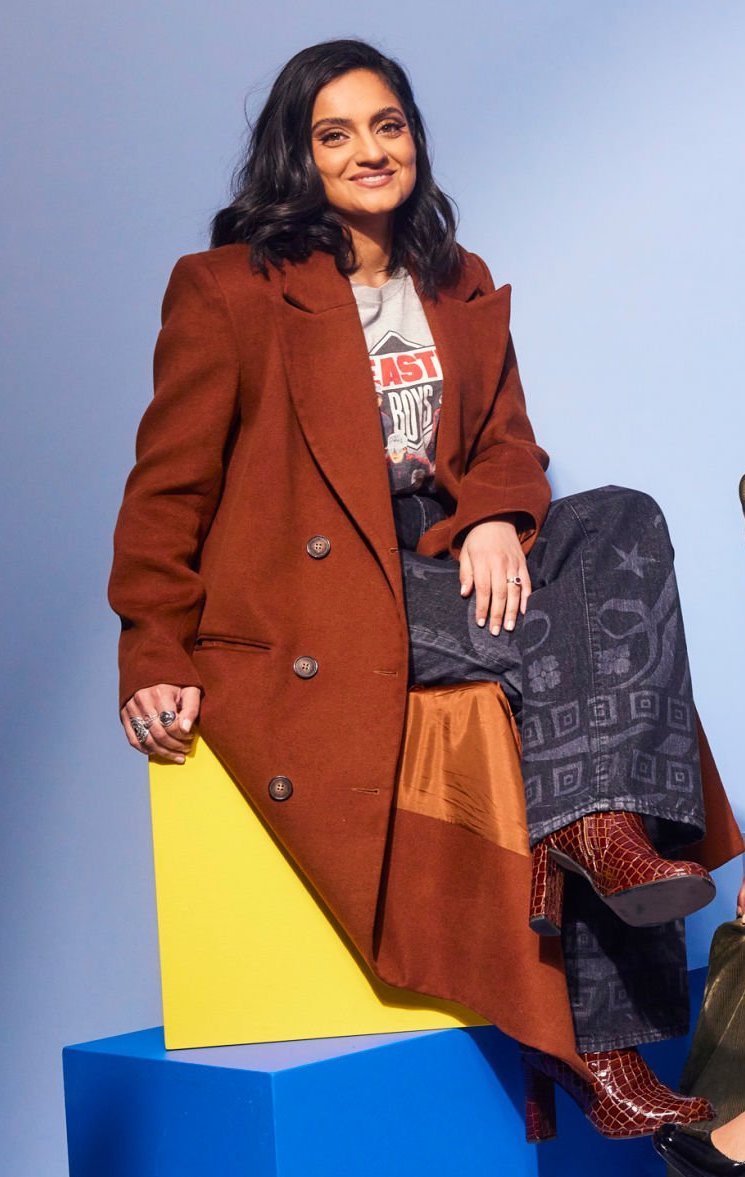 Portrait of a smiling woman in a rust-colored coat sitting on blue and yellow blocks against a blue background in a studio.