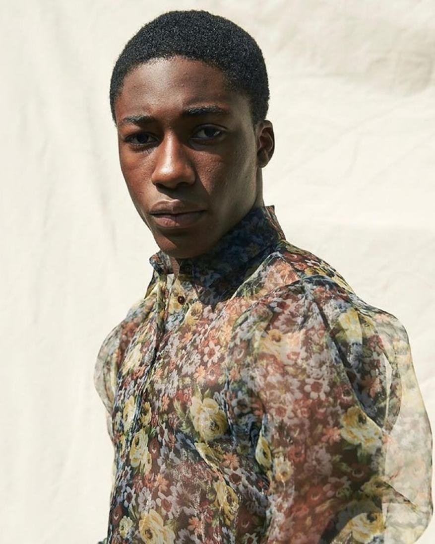 Close-up portrait of a Black man with short hair wearing a sheer floral patterned shirt against a neutral backdrop.