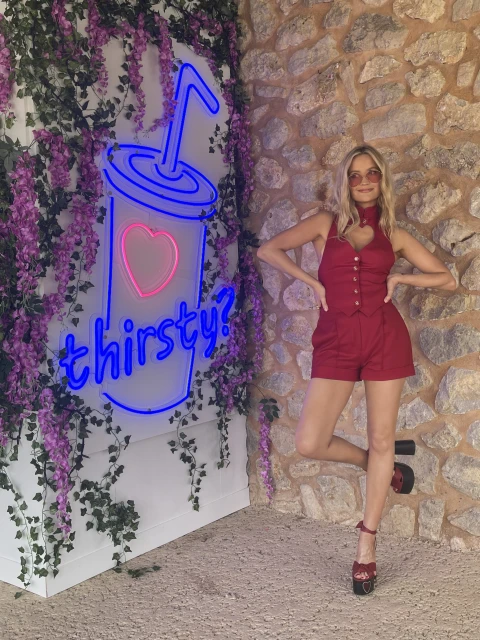 Blonde woman poses in a red outfit next to a neon sign that says 'thirsty?' with lush greenery and a stone wall backdrop.