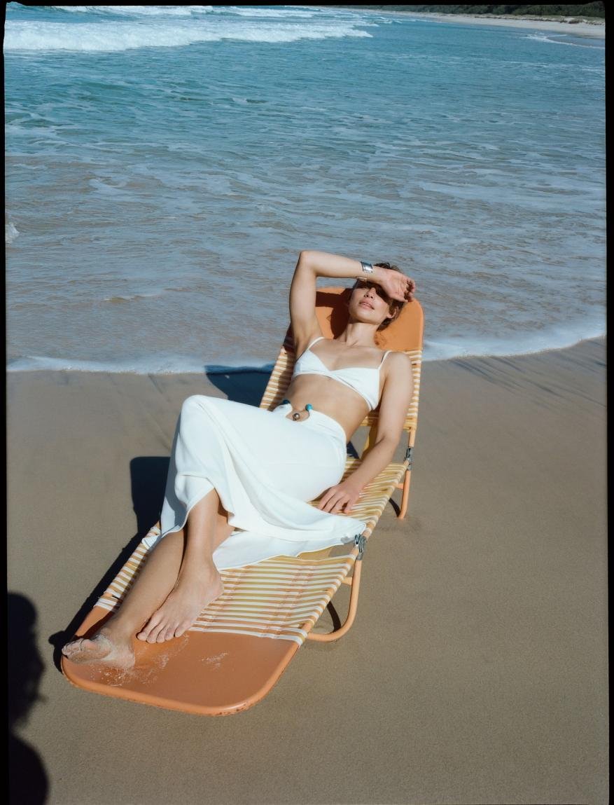 Woman in a white bikini and sarong lounges on an orange beach chair on a sandy beach near the ocean, shielding her eyes from the sun.
