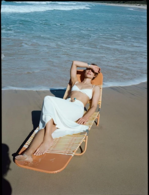 Woman in a white bikini and sarong lounges on an orange beach chair on a sandy beach near the ocean, shielding her eyes from the sun.