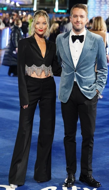 A stylish couple poses on a blue carpet at a formal event. The woman wears a black suit with beaded trim, and the man wears a blue velvet suit.