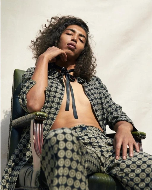 Male fashion model with curly hair poses in patterned pants and jacket, seated in a green chair in an indoor setting with natural light.